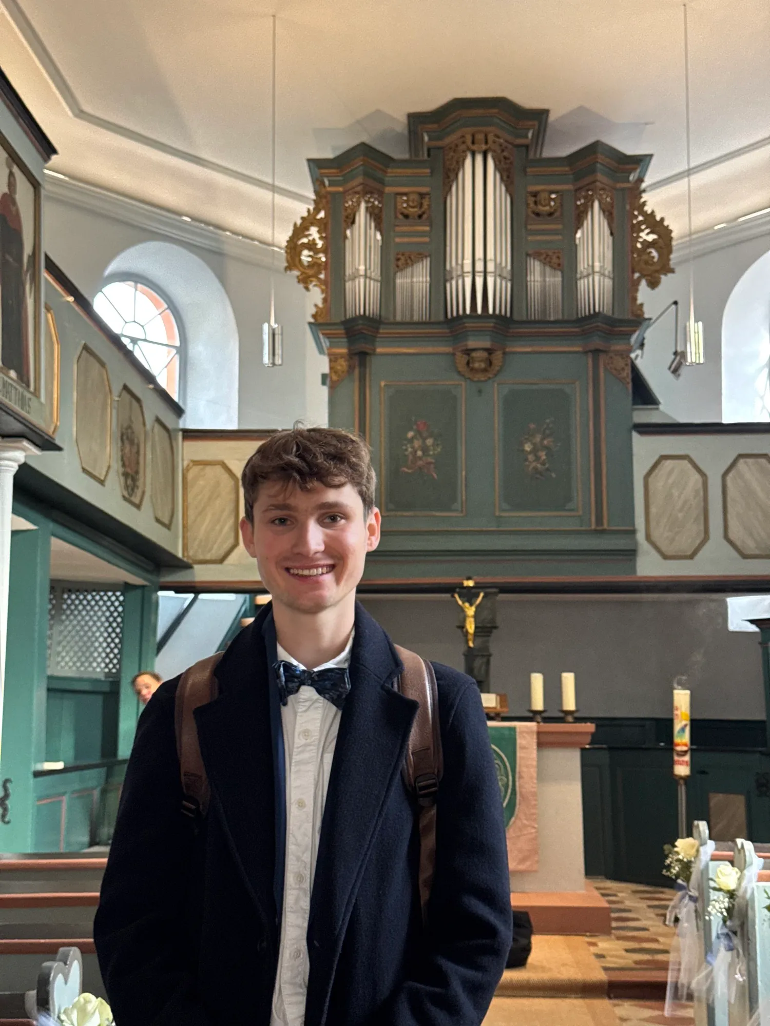 Lukas Höver in a coat and bow tie in front of a church organ case, with flowers on the pews behind him